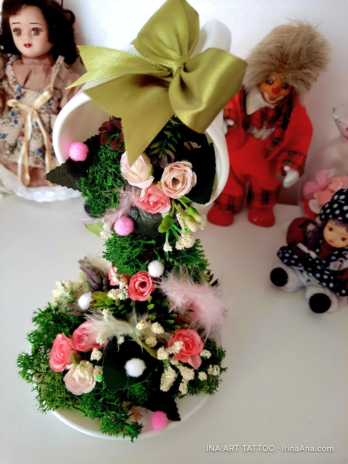 A white teacup, overturned, with a cascade of pink and green flowers spilling out, adorned with a golden ribbon.