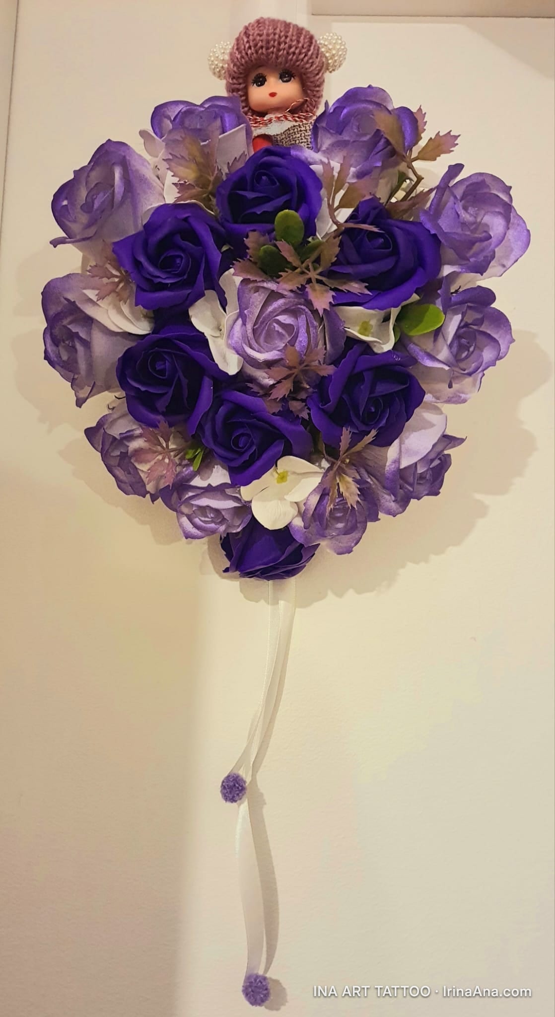 Decorative bouquet with purple artificial roses and a small doll with a knitted hat, hanging on a white wall.
