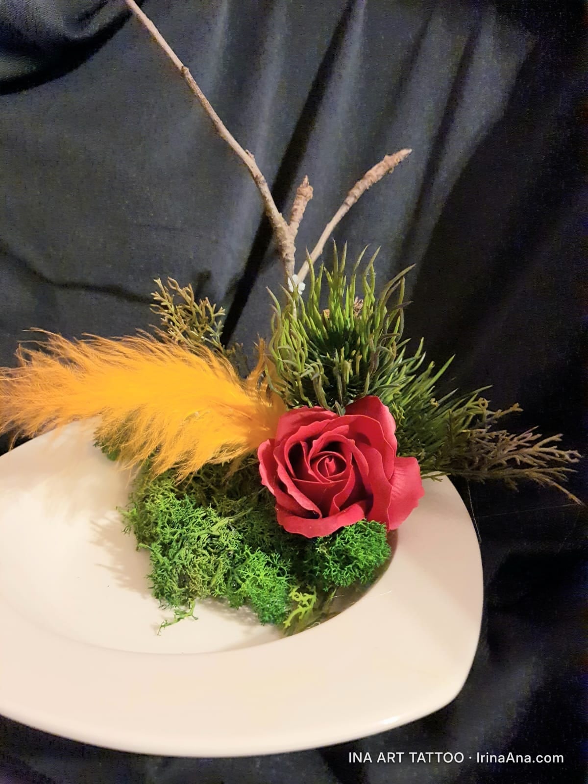 Handmade floral arrangement with a red crepe paper rose, green moss, a yellow feather, and dried twigs, on a white plate.