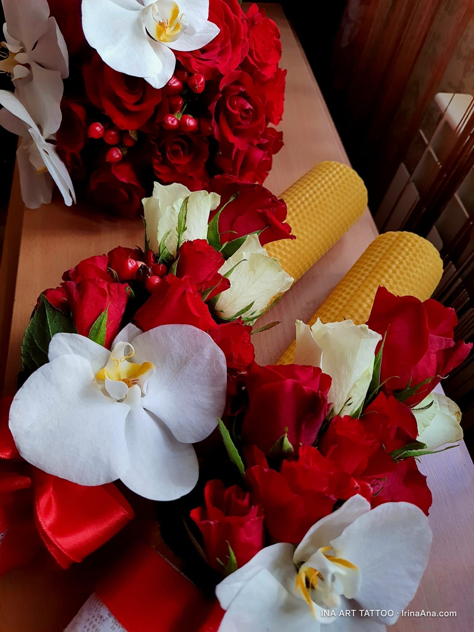Two artificial flower bouquets, red and white, with orchids and roses, next to two yellow candles.