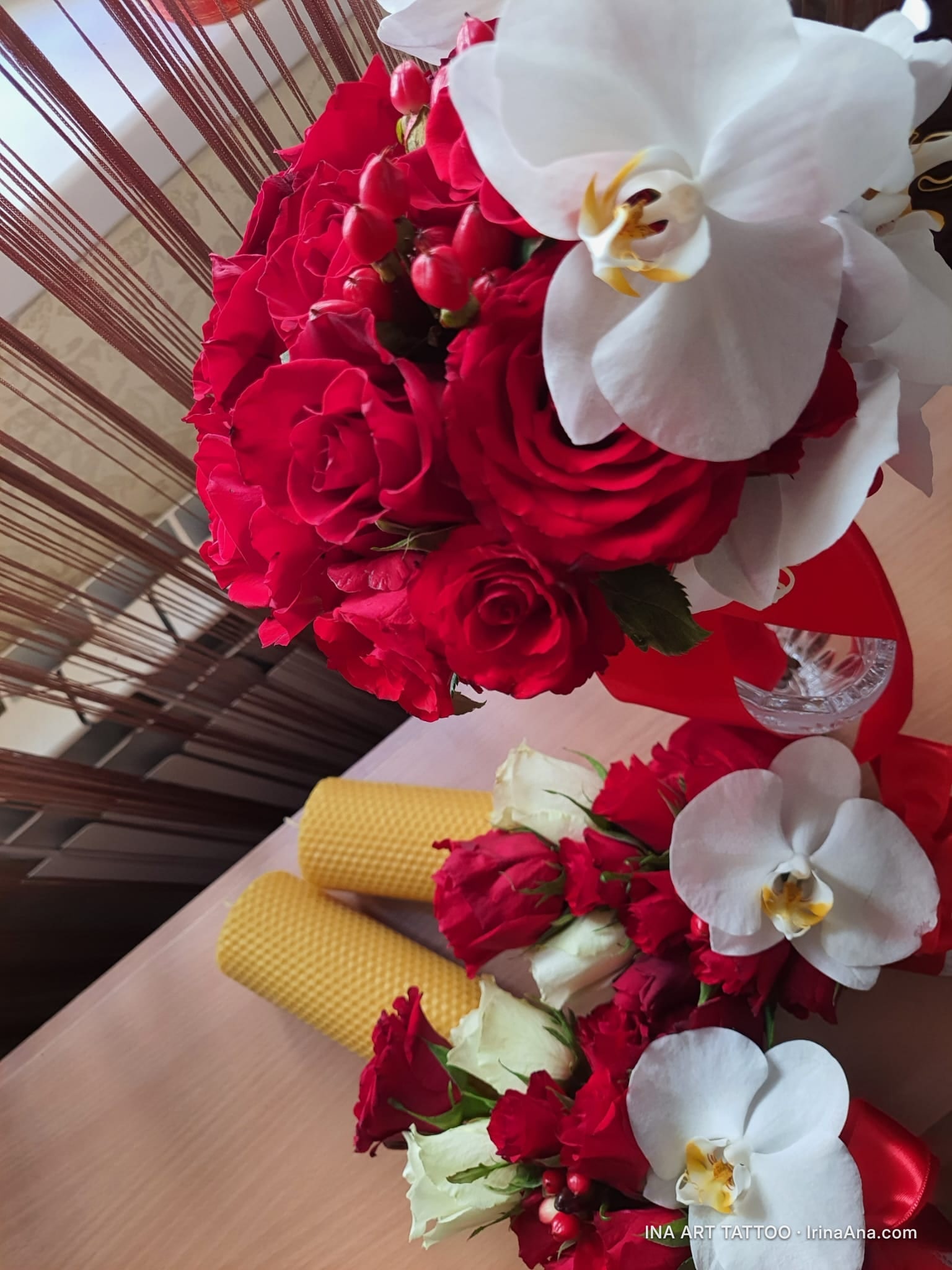 Two bouquets of red roses, white orchids, and red berries, next to two yellow wax candles.