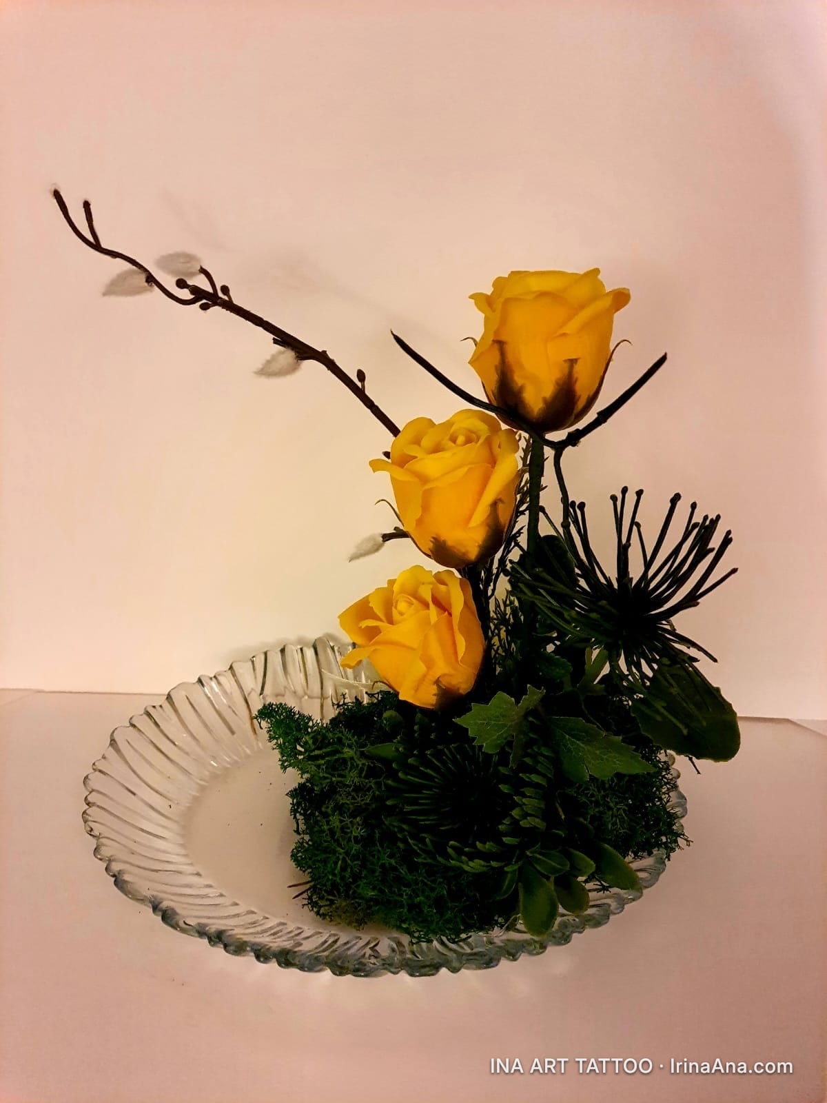 Three yellow crepe paper roses arranged with green moss and dry branches in a glass dish.