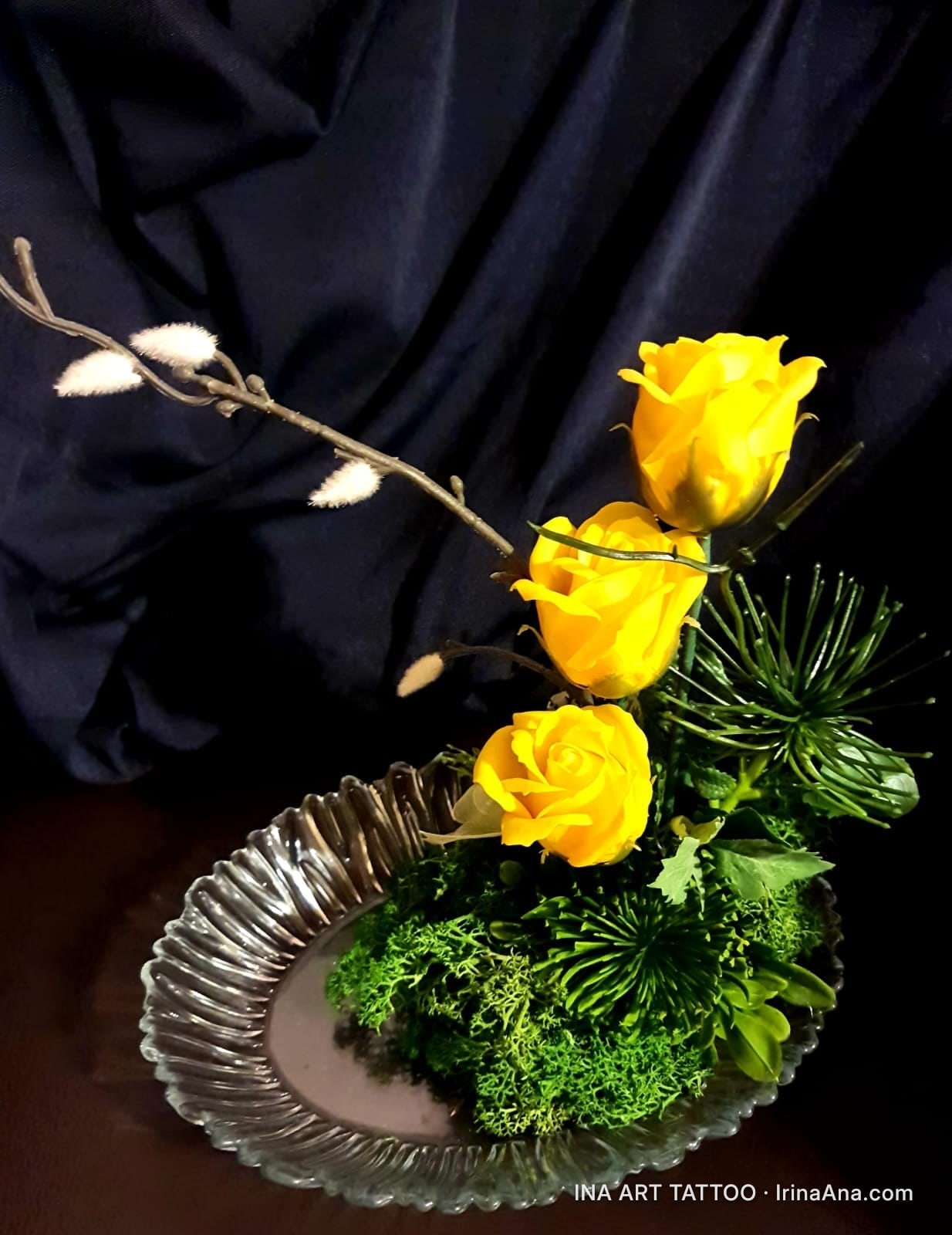 Floral arrangement with three yellow roses, green moss, and twigs, in an oval glass dish, against a black background.
