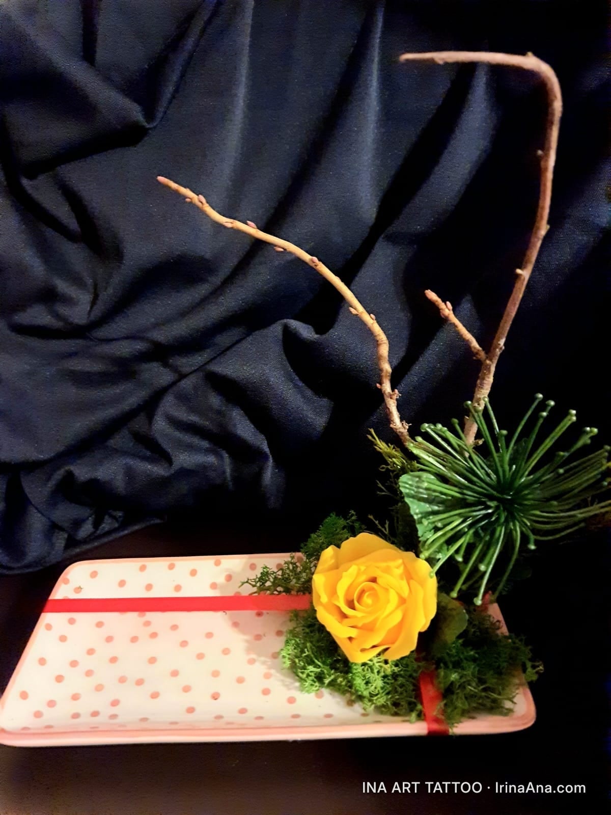 Handmade floral arrangement with a yellow rose, green moss, twigs, and a decorative plant on a white tray with pink polka do…
