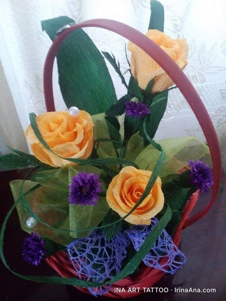 A decorative basket with yellow crepe paper roses, purple flowers, and green ribbons.