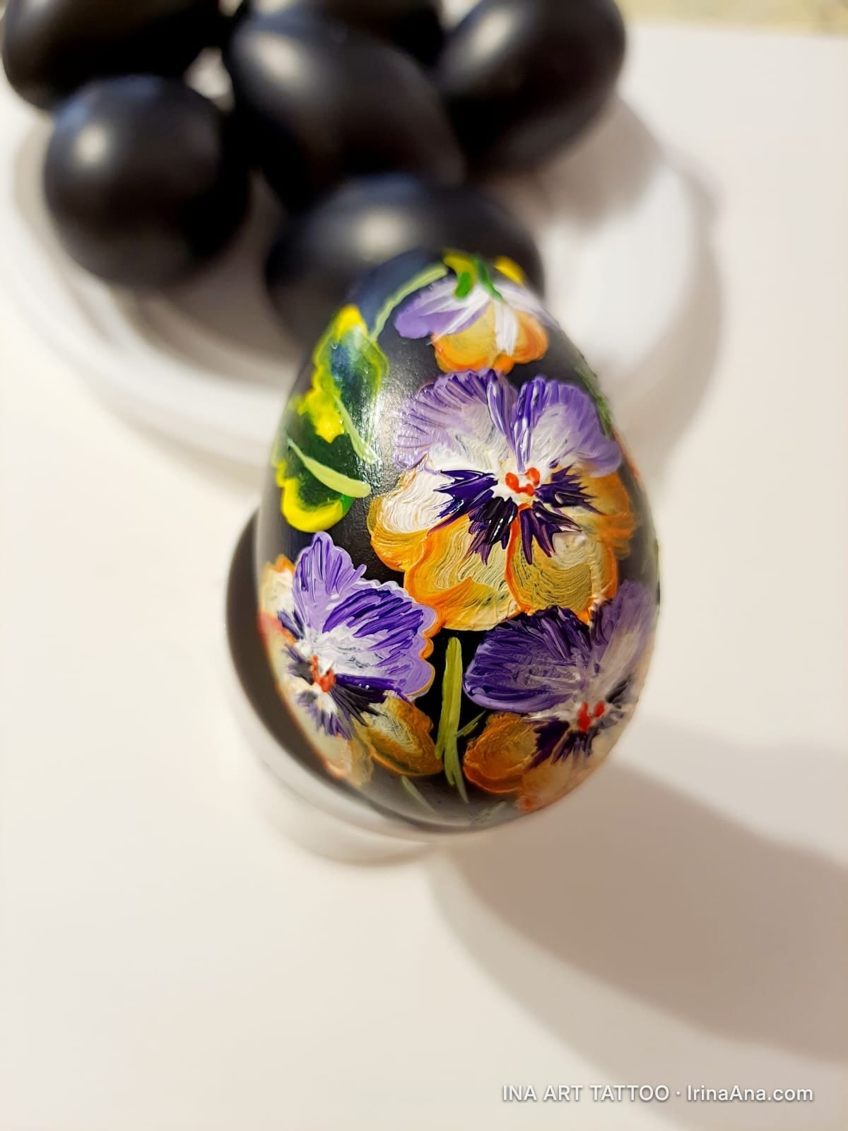 A black Easter egg, hand-painted with pansy flowers in shades of purple, yellow, and orange. Other black eggs are in the bac…