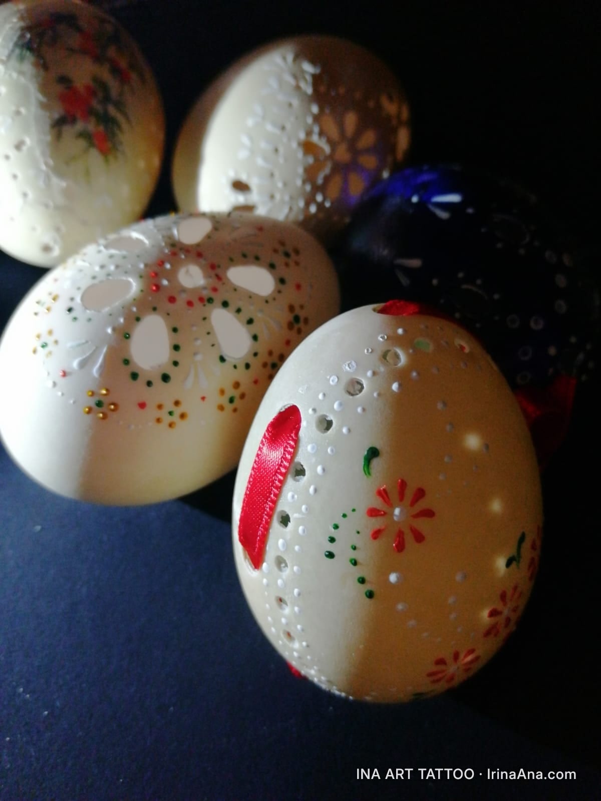 Five decorated eggs, three white with perforated and painted floral patterns, one with red flowers, one black. Dark backgrou…