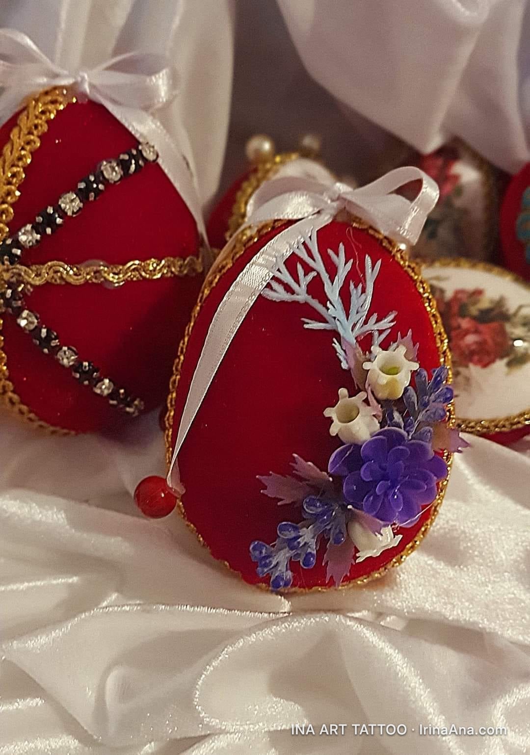 A red velvet decorative egg, with a white bow and small blue, violet, and white flowers, on a white cloth.