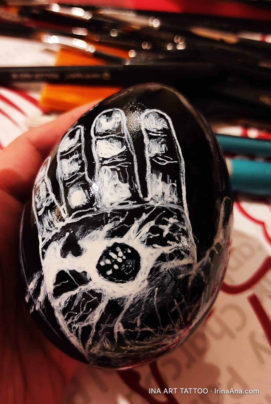 A hand holds a black egg, engraved with a white, skeletal hand holding a small skull. Blurred background with paintbrushes.