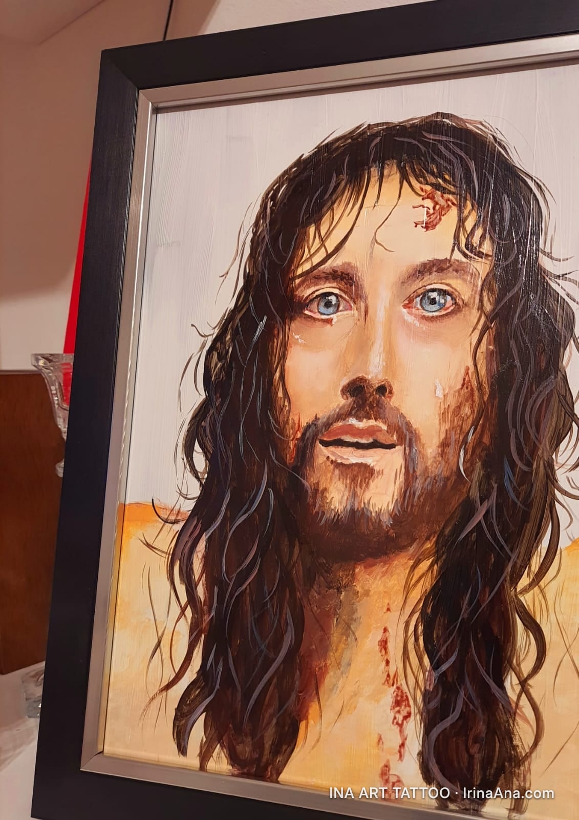 Painted portrait of a man with long hair, beard, blue eyes, and blood marks on his face and chest. Black frame.