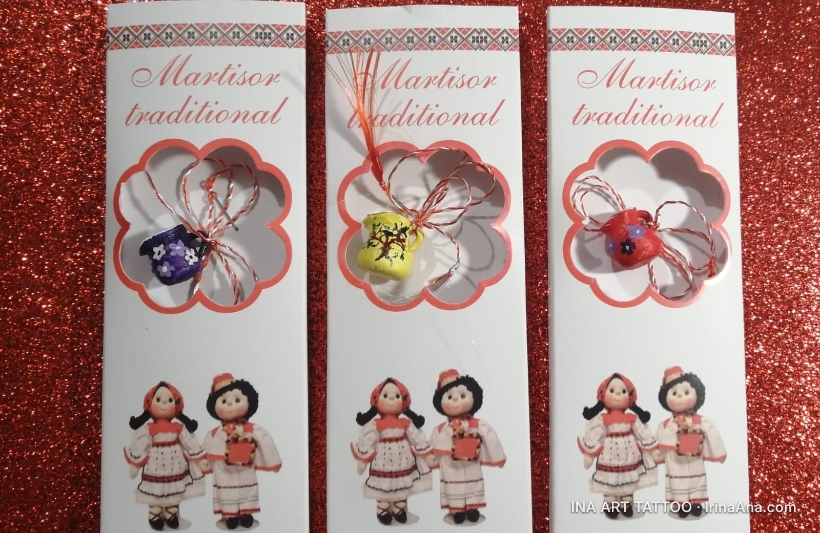 Three traditional Martisor cards, each with a folk doll and a small pitcher-shaped pendant, on a glittery red background.