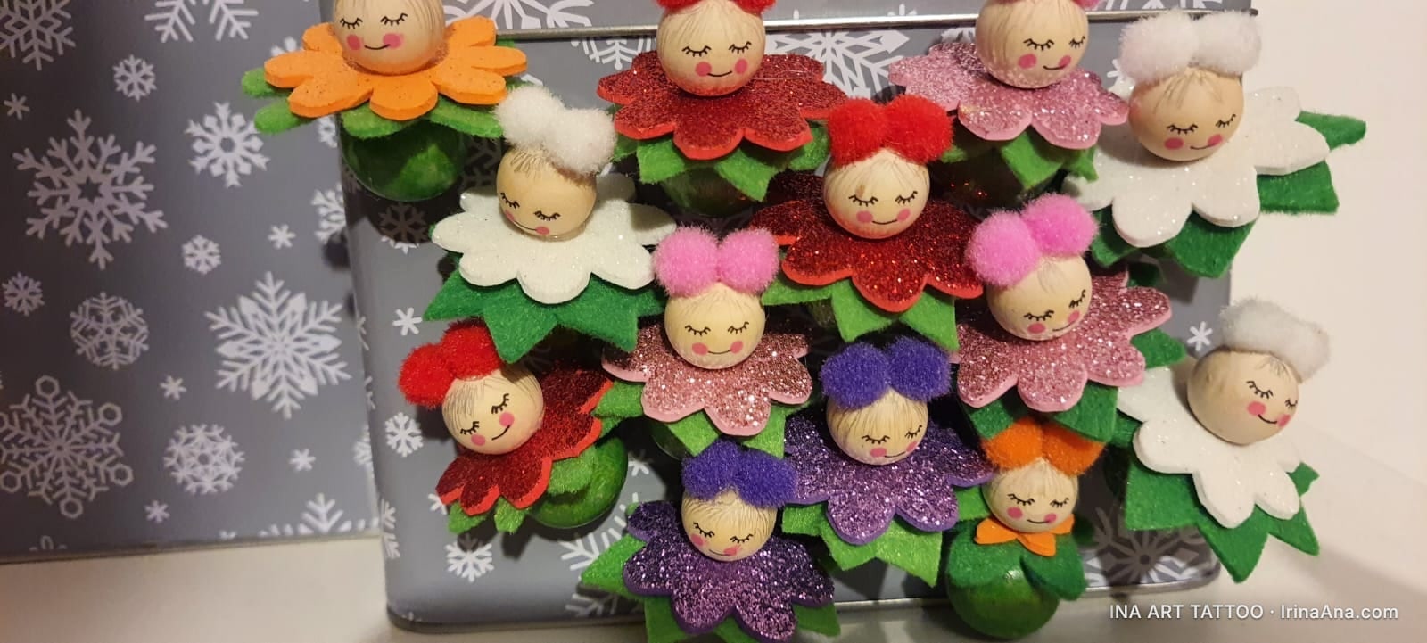 Eleven handmade felt dolls with smiling faces and colorful dresses, against a snowflake-patterned background.