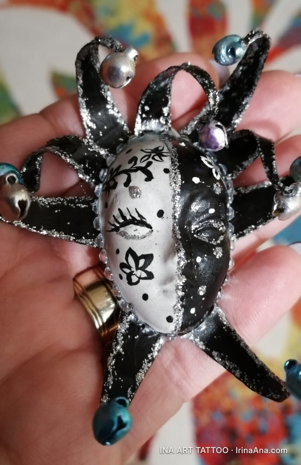A black and white harlequin mask, with silver glitter and bells, held in a palm.