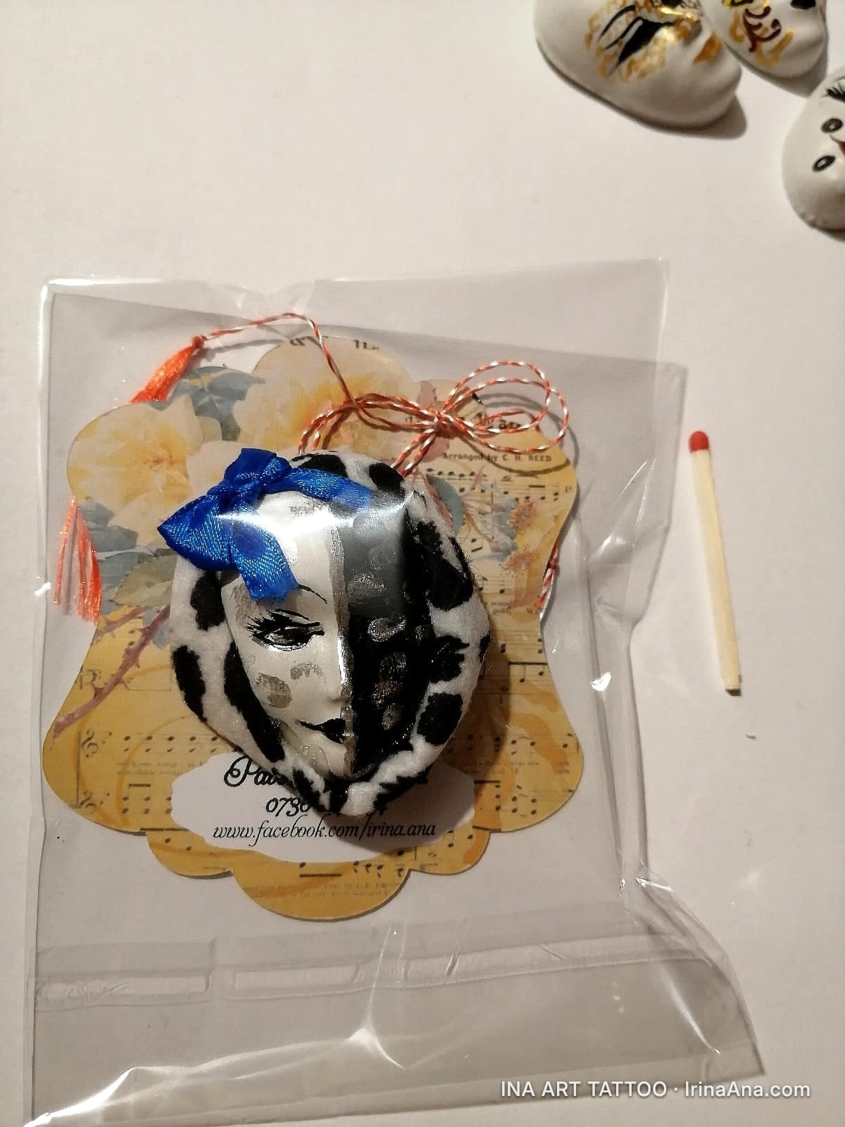Mărțișor in the shape of a harlequin mask, half white with black spots, half black with white spots, with a blue bow.