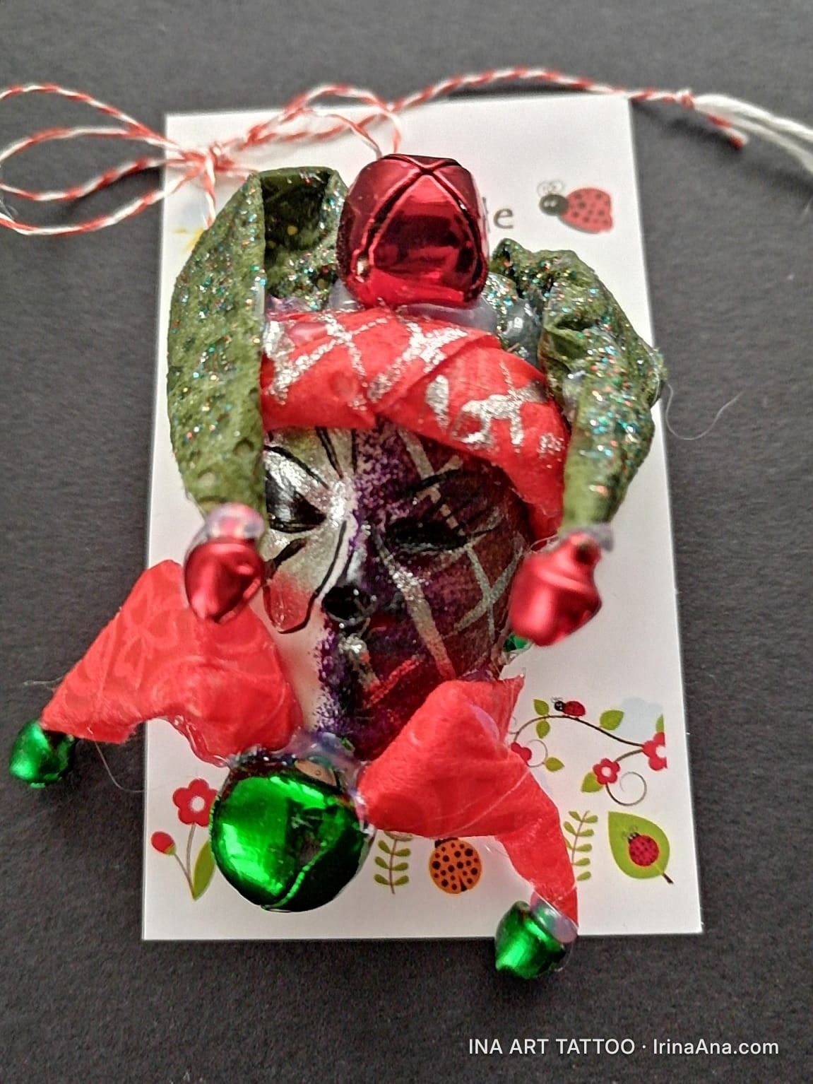 Mărțișor in the shape of a harlequin mask, with red and green ribbons, red and green bells, on a white card.