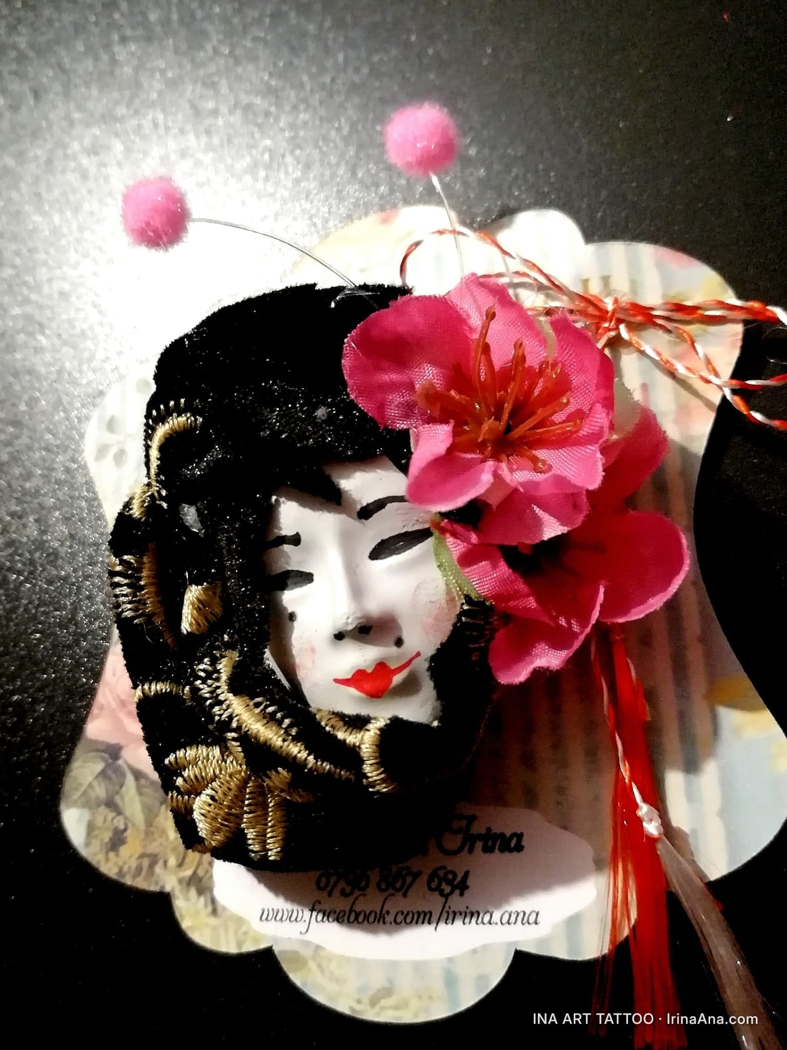 Handmade mărțișor with a white Japanese mask, black and gold embroidery, a pink flower, and red and white tassels.