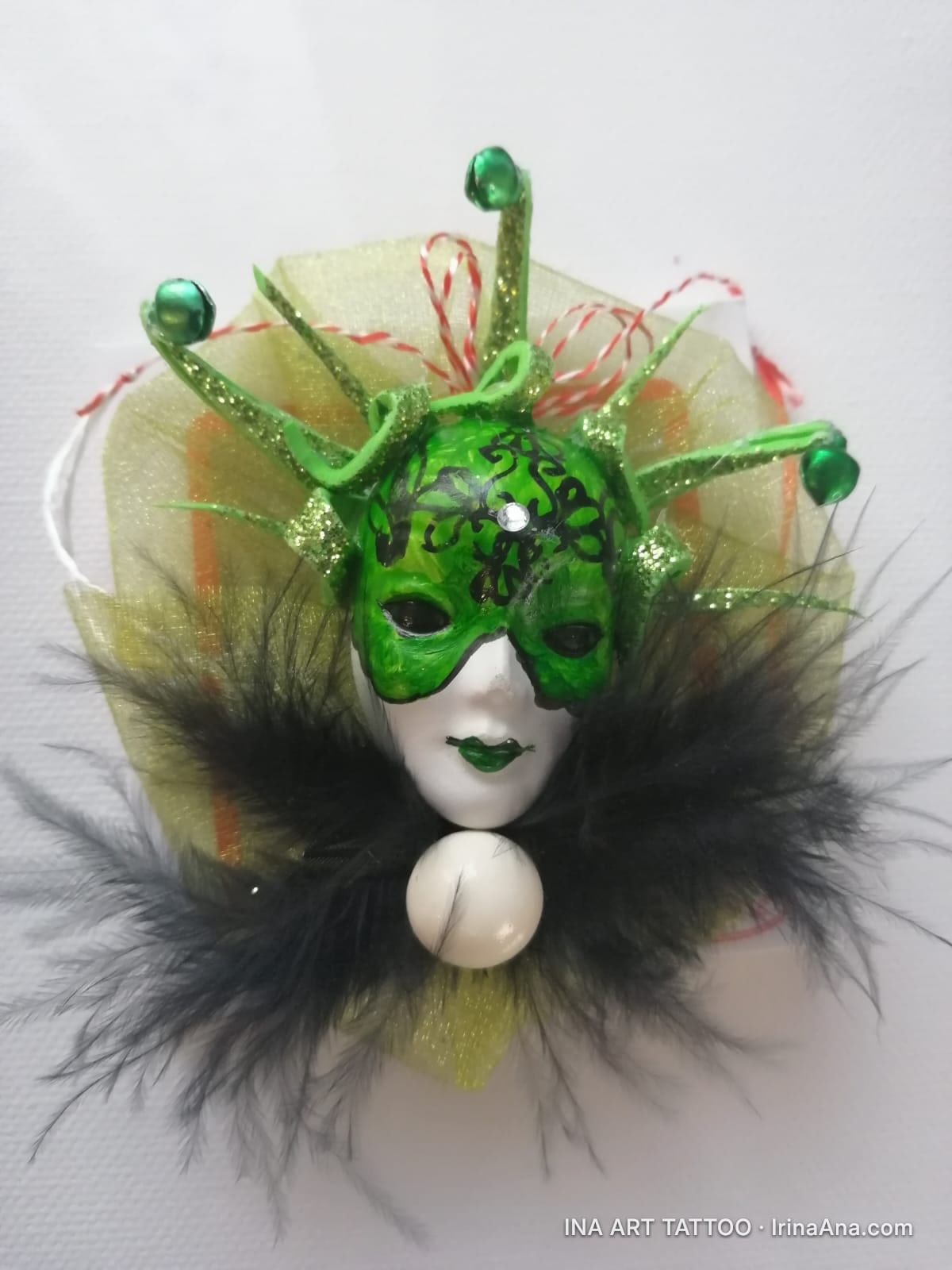 A mărțișor in the shape of a carnival mask, with a green mask, black feathers, and a white bead under the chin.