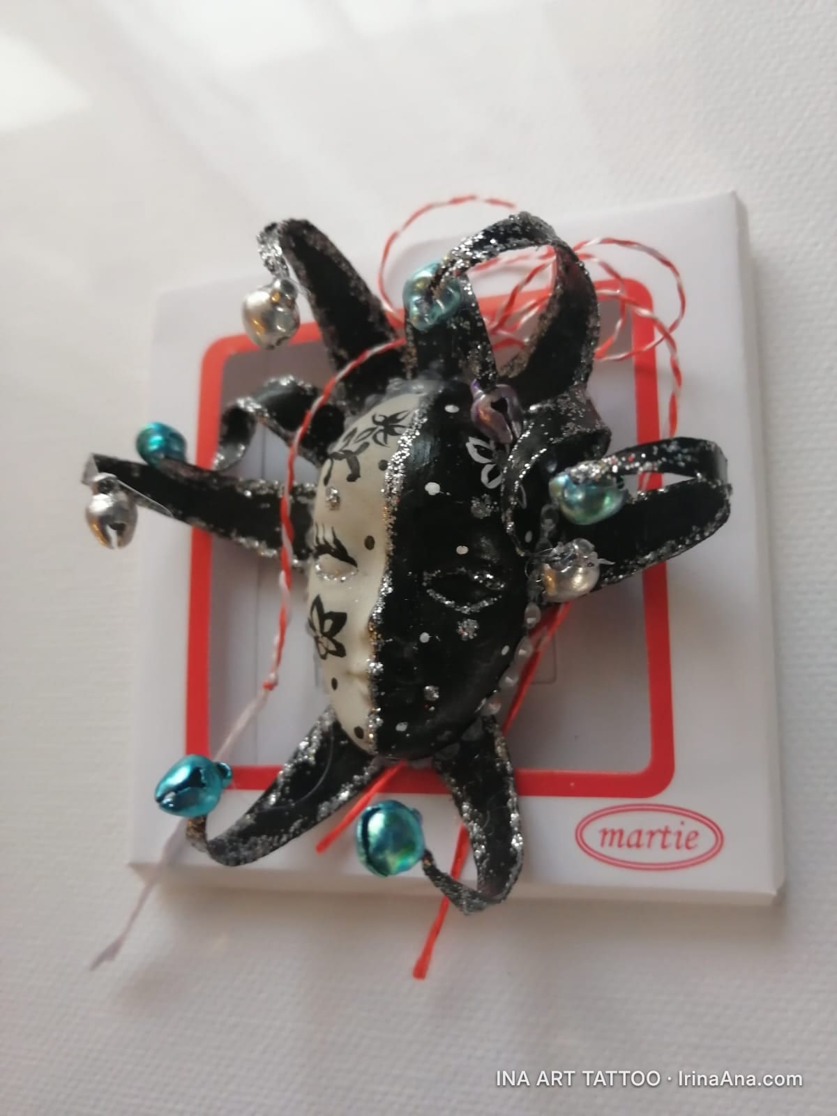Martisor shaped like a jester mask, half white with black flowers, half black with white polka dots, with bells.