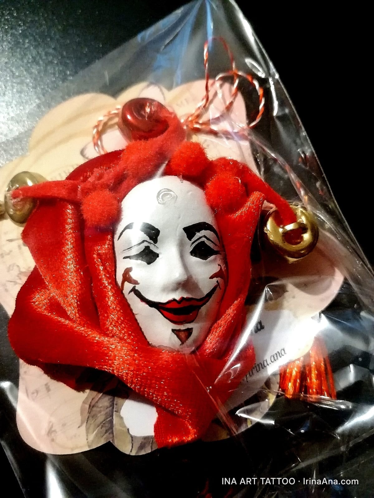 A martisor featuring a white jester mask, surrounded by red fabric and golden bells, packaged in clear plastic.