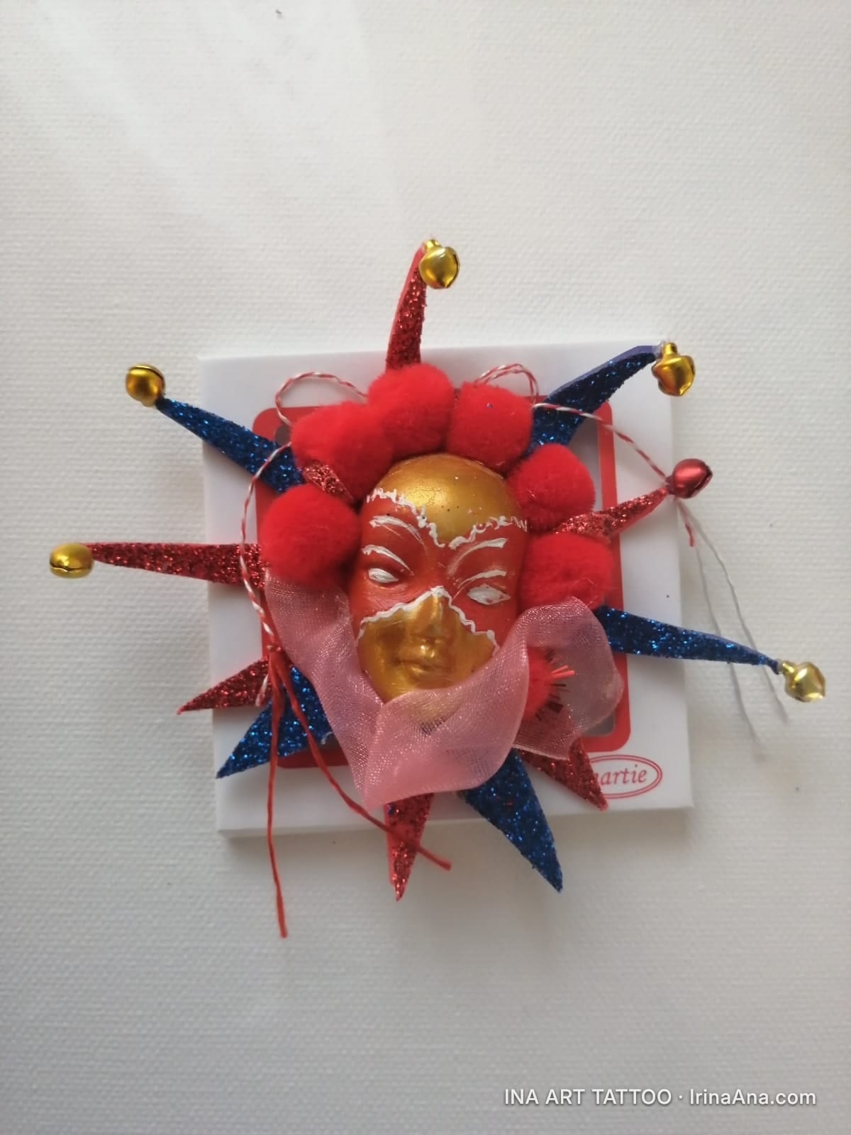 Mărțișor: red and gold jester mask with pointed red and blue collar, decorated with pom-poms and golden bells.