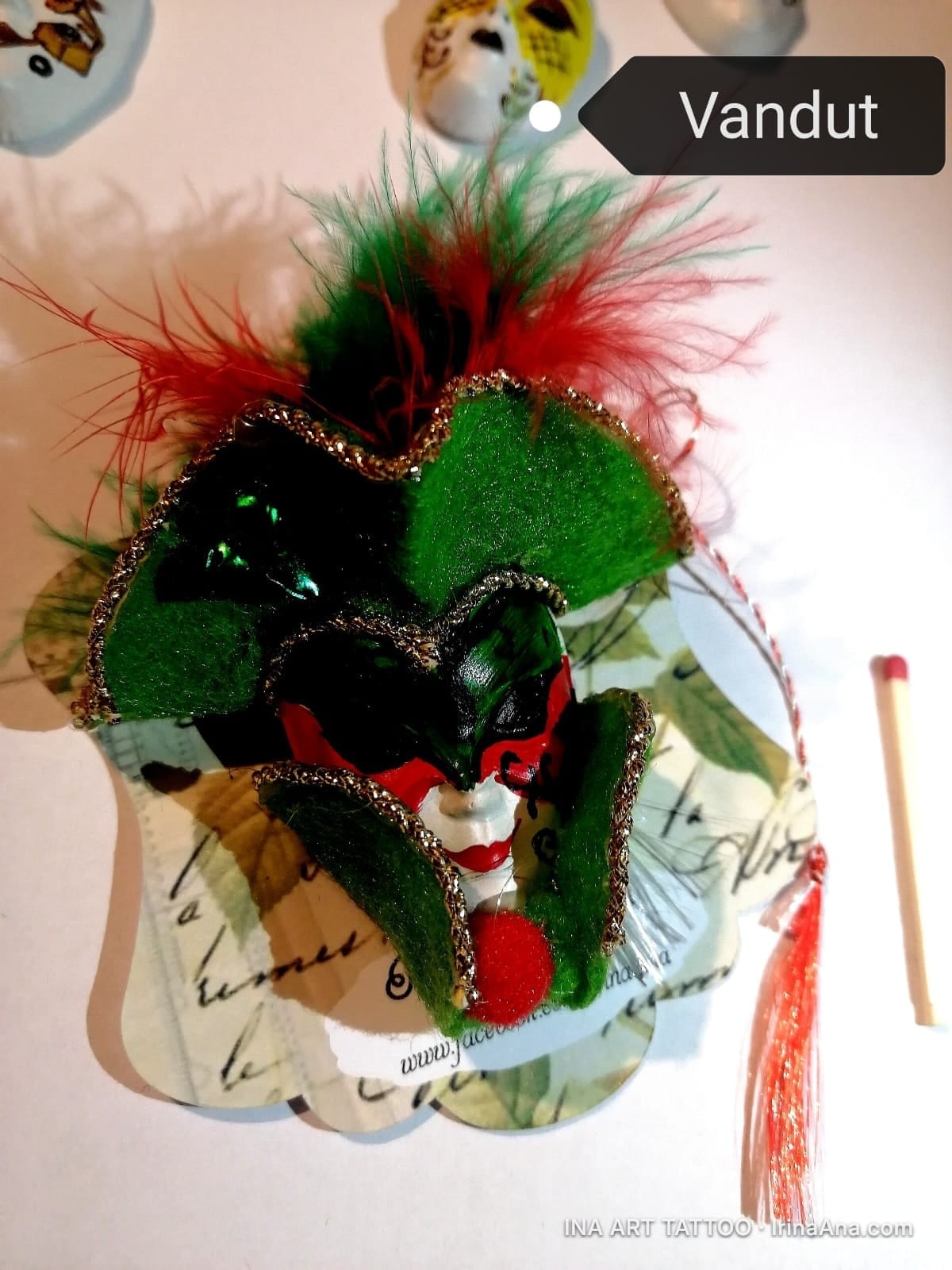 A jester mask shaped mărțișor, with red and green feathers, gold details, and a white face with red lips.
