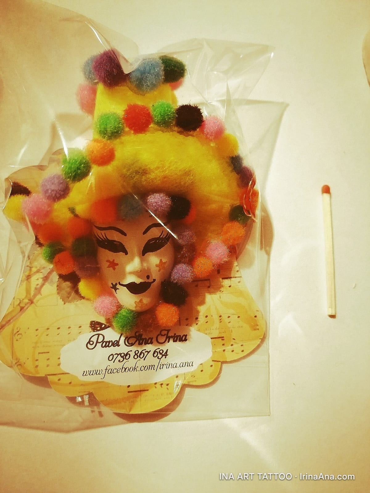 A Mardi Gras mask with fluffy yellow hair and colorful pom-poms, packaged in a clear plastic bag, next to a matchstick.