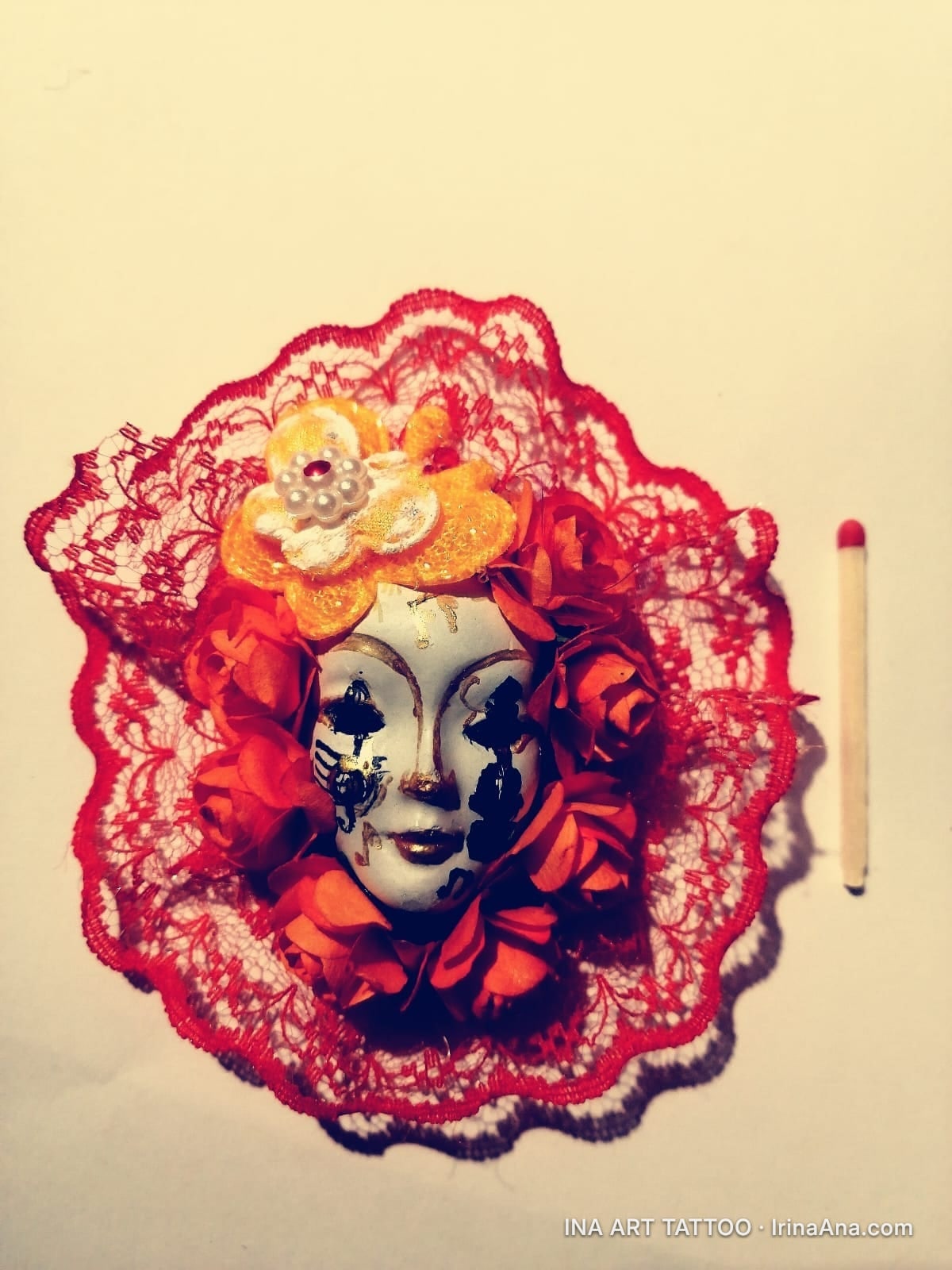Mărțișor: white mask with black and gold spots, surrounded by red-orange flowers and red lace, next to a matchstick.