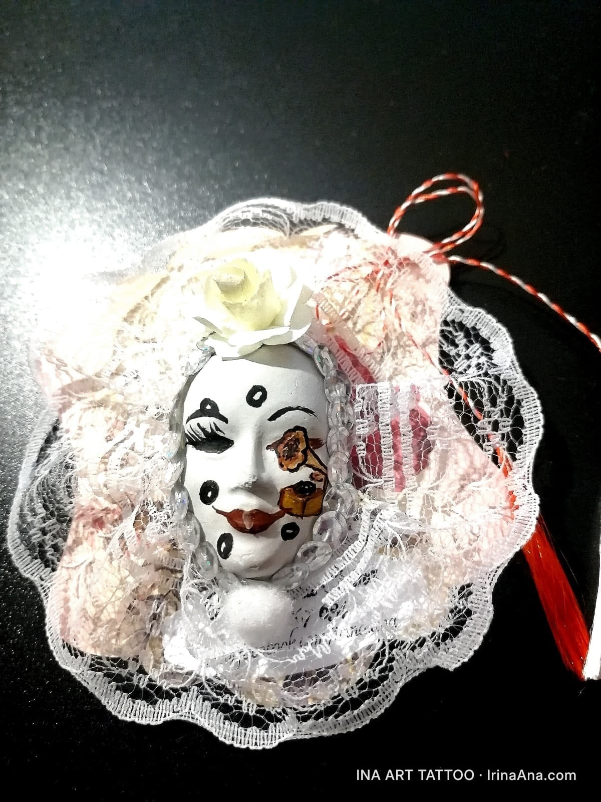 Mărțișor: white ceramic mask with clown makeup, surrounded by white lace and red and white ribbons.