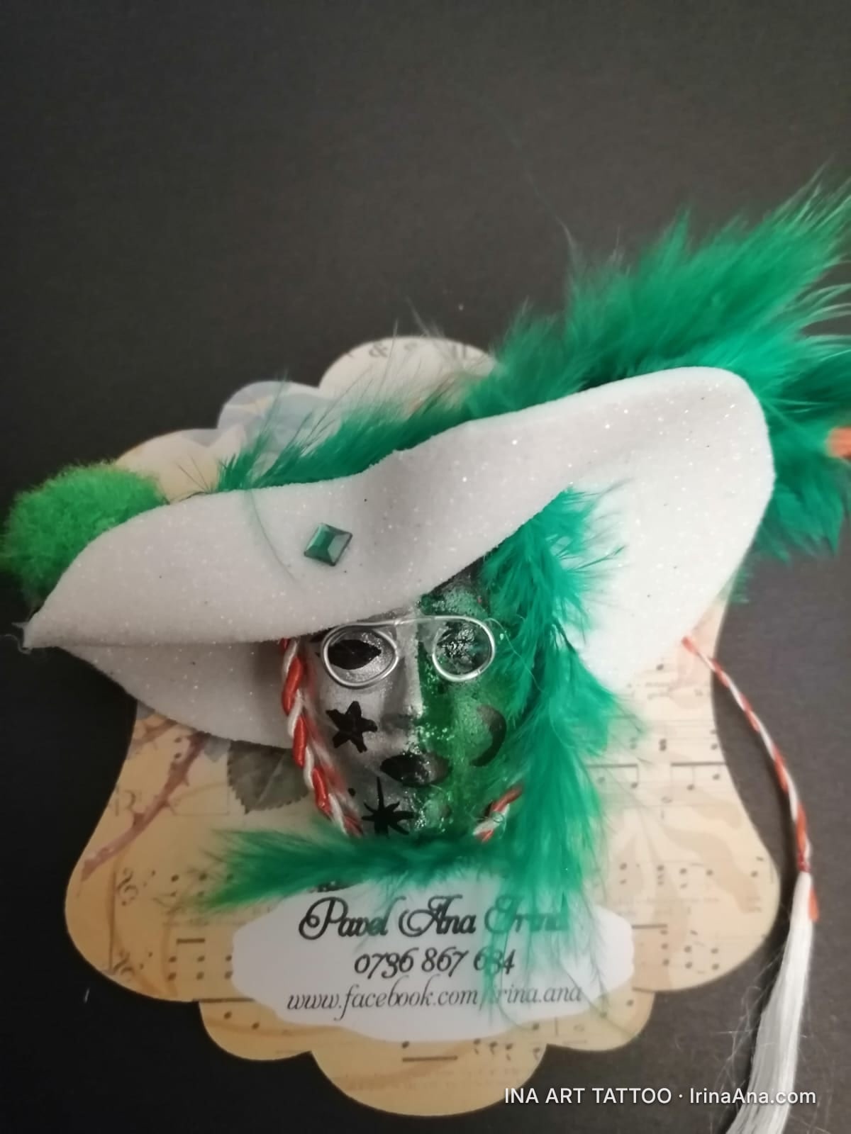 A handmade mărțișor featuring a green mask with a white hat decorated with green feathers and an orange feather.
