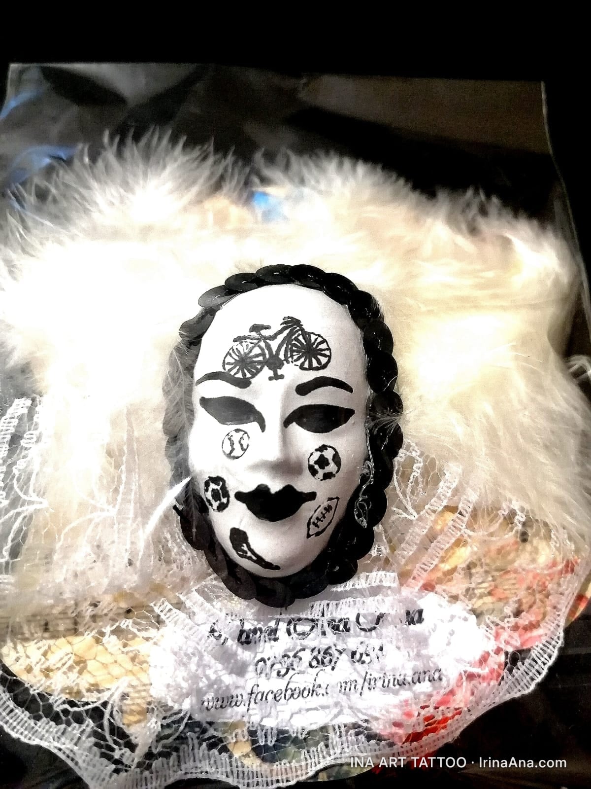 A white mask with a bicycle on the forehead and sports symbols on the cheeks, surrounded by white feathers and lace.