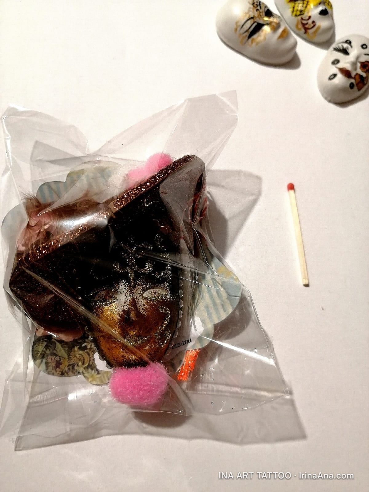 Handmade Mărțișor craft object, a mask decorated with glitter and pink pompom, in a clear bag. Next to it, a matchstick and…