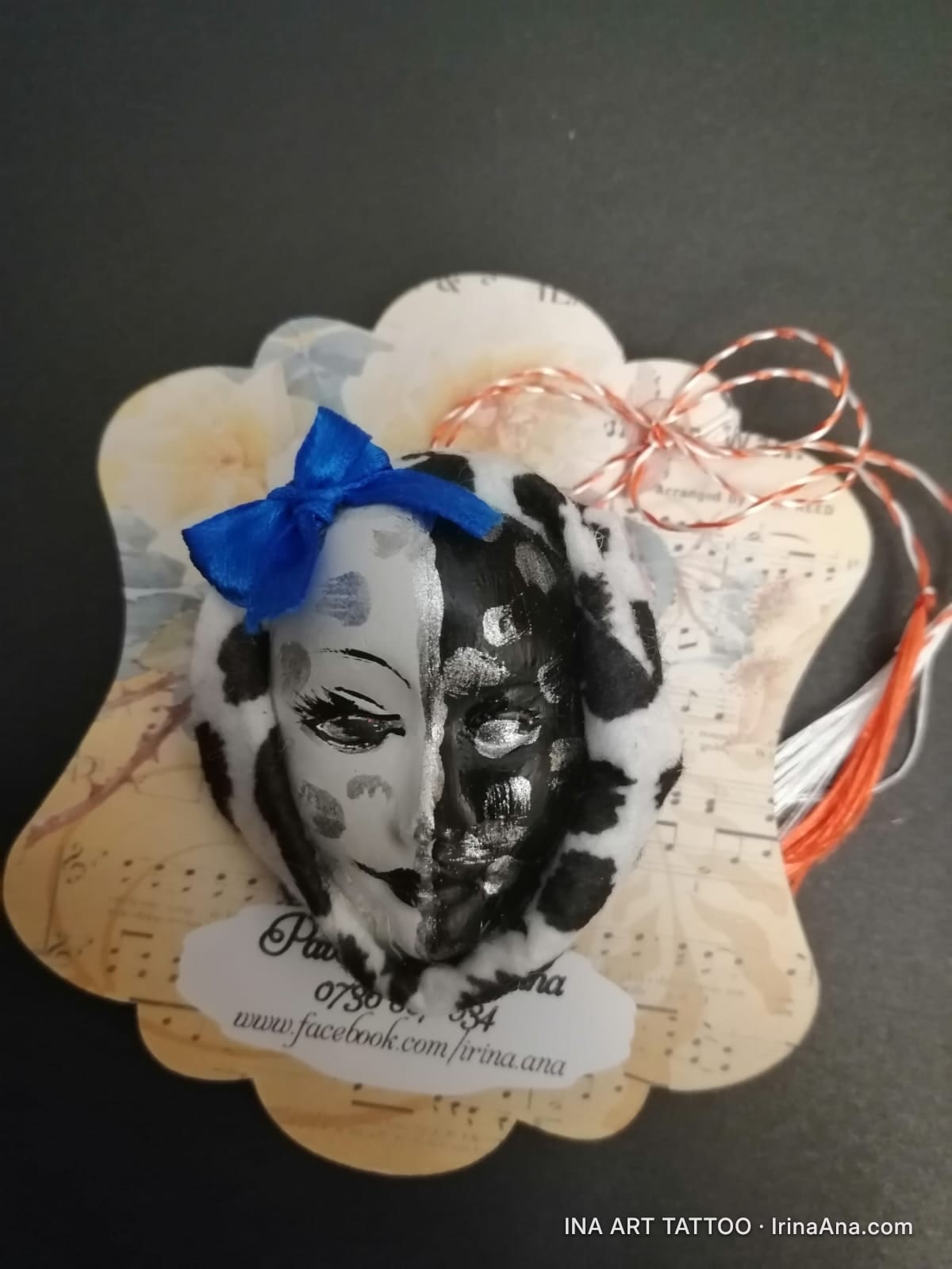 Mărțișor: black and white polka dot mask, one half silver, blue bow, on a cardboard backing with musical notes and string.
