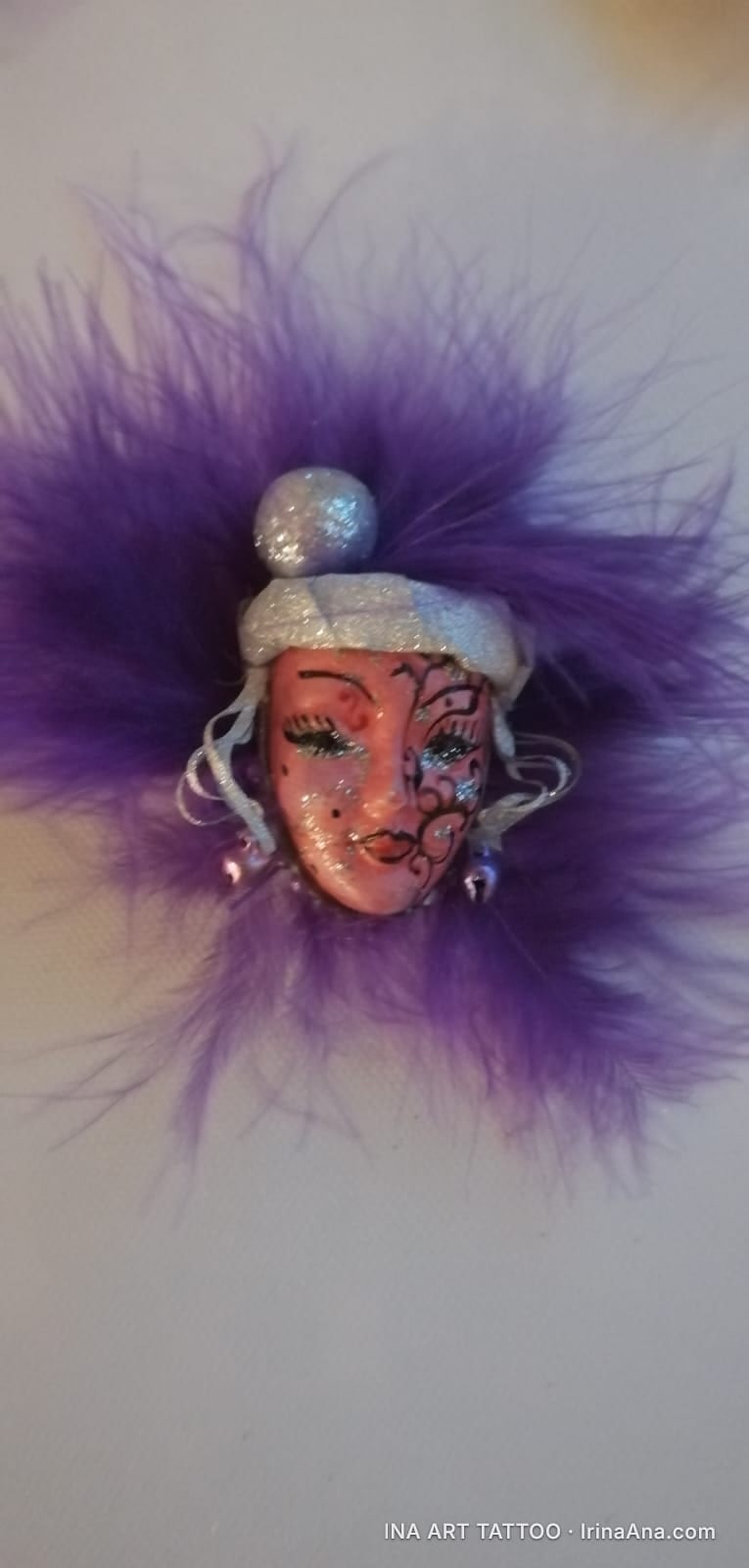 Mărțișor in the shape of a decorative mask with elaborate makeup and purple feathers, on a white background.