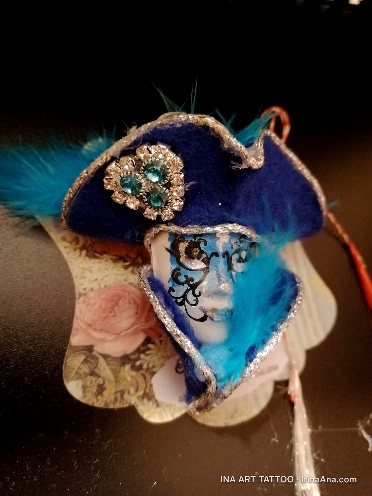 Mărțișor: a blue Venetian mask with a hat and feathers, decorated with shiny stones. Black background.