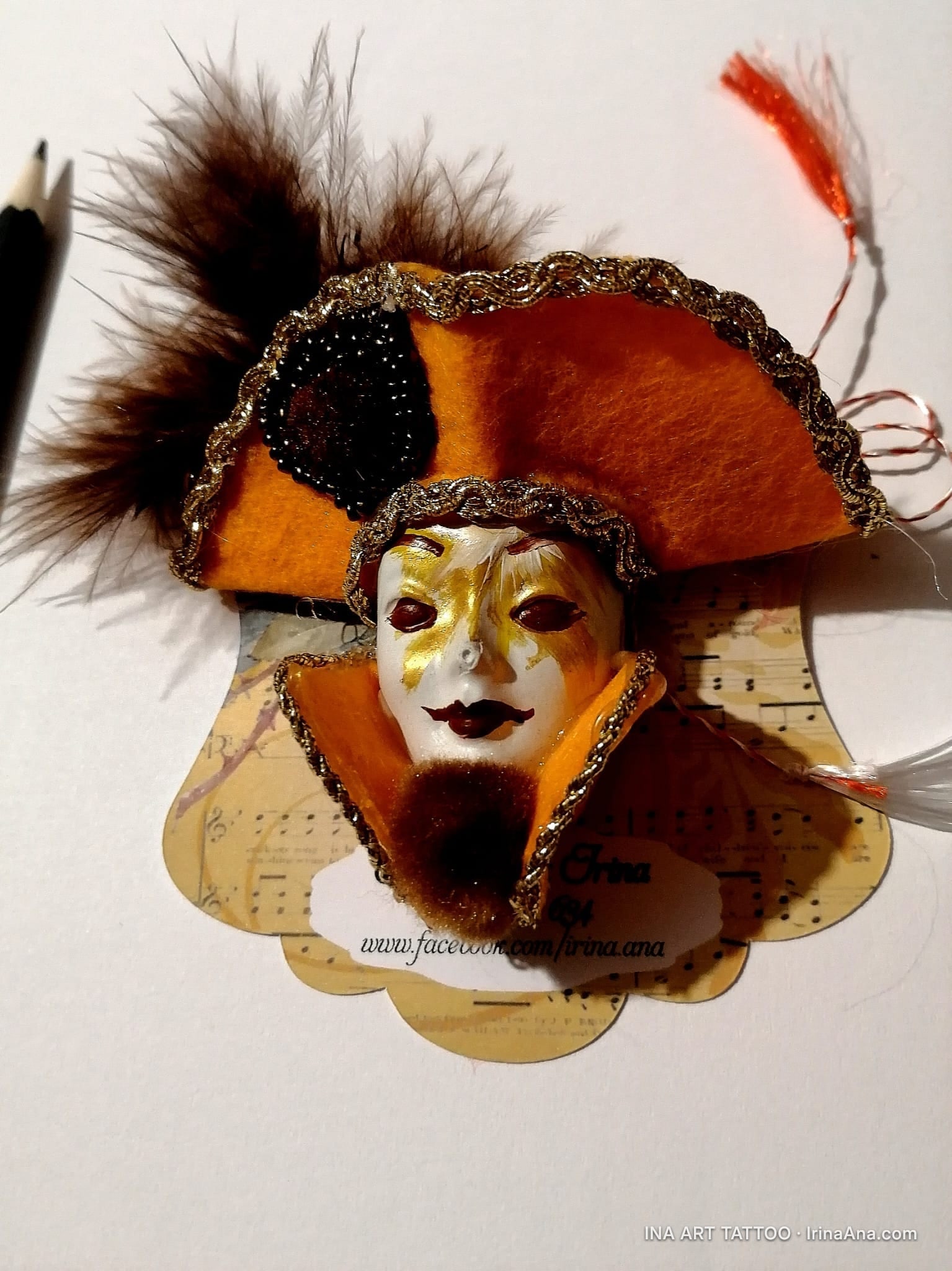 Martisor brooch shaped like a Venetian mask, with an orange hat, brown feathers, and gold details, on a white background.