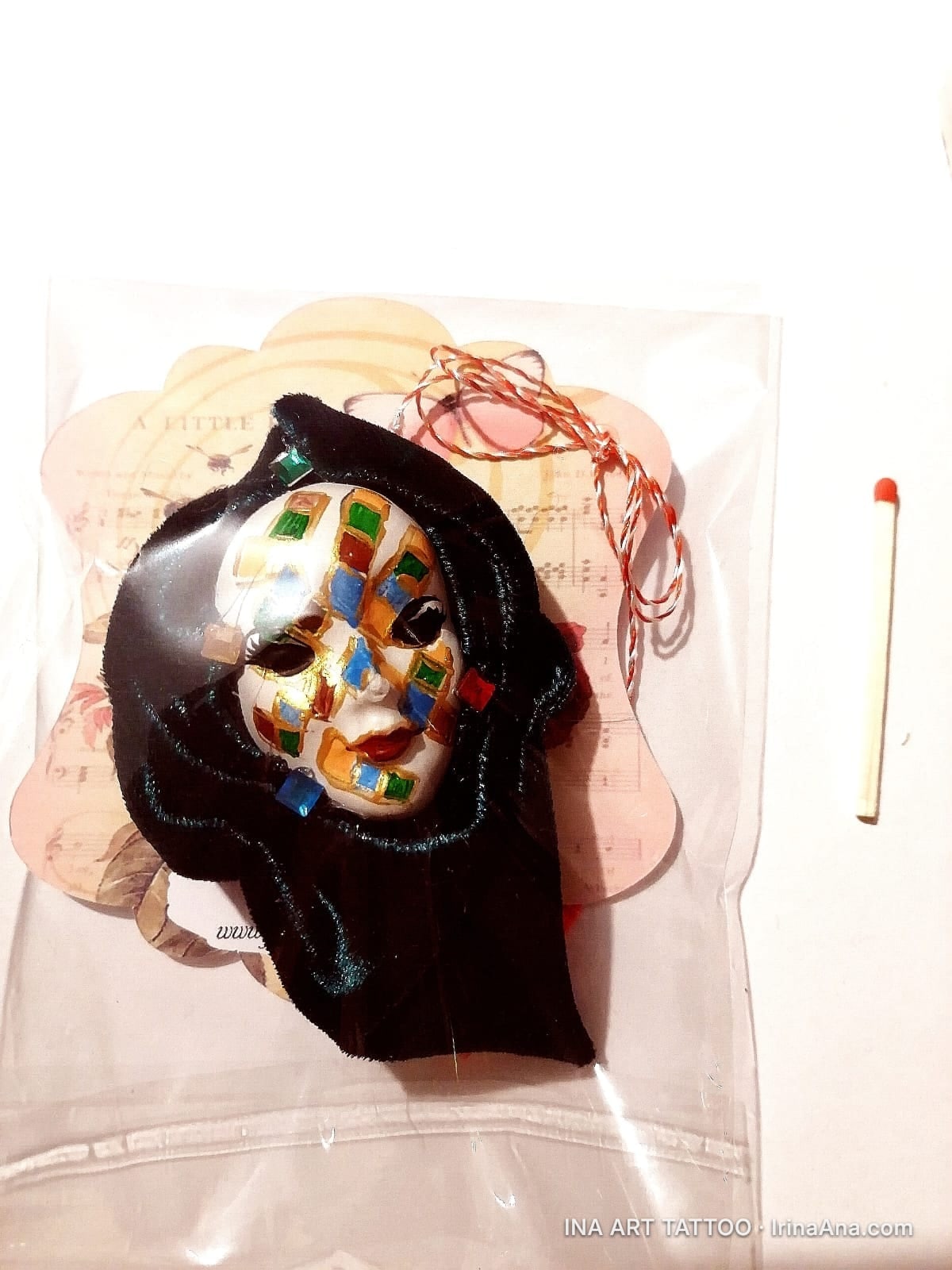 Martisor brooch of a white Venetian mask with colorful patches and a black hood, packaged in a transparent bag.