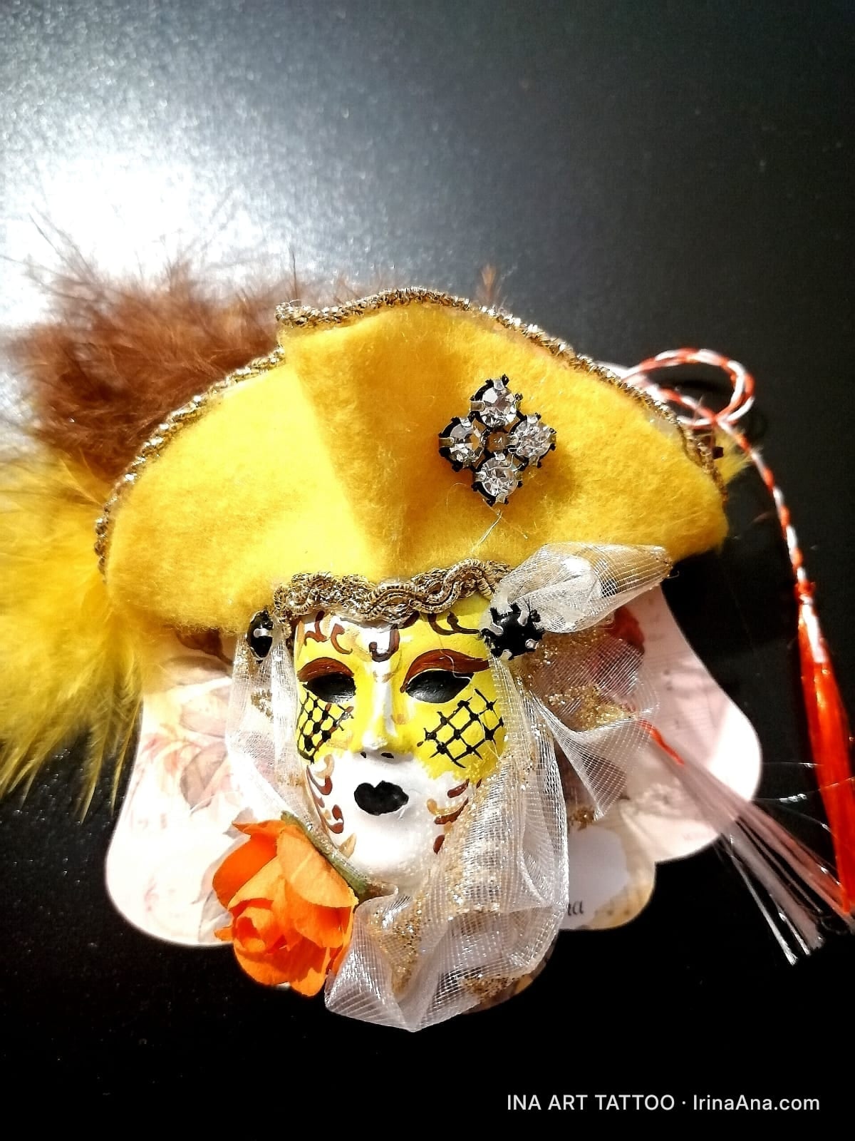 Martisor brooch: white and yellow Venetian mask with a yellow hat, brown feathers, an orange rose, and a red-white string.