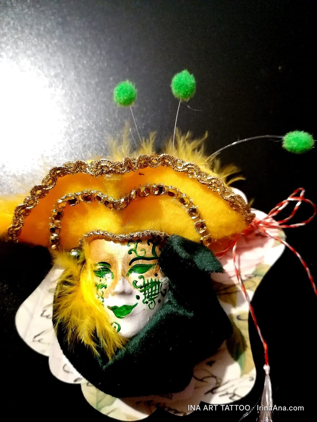 Mărțișor: white Venetian mask with green and gold details, yellow hat with feathers, green pompoms, and red string.