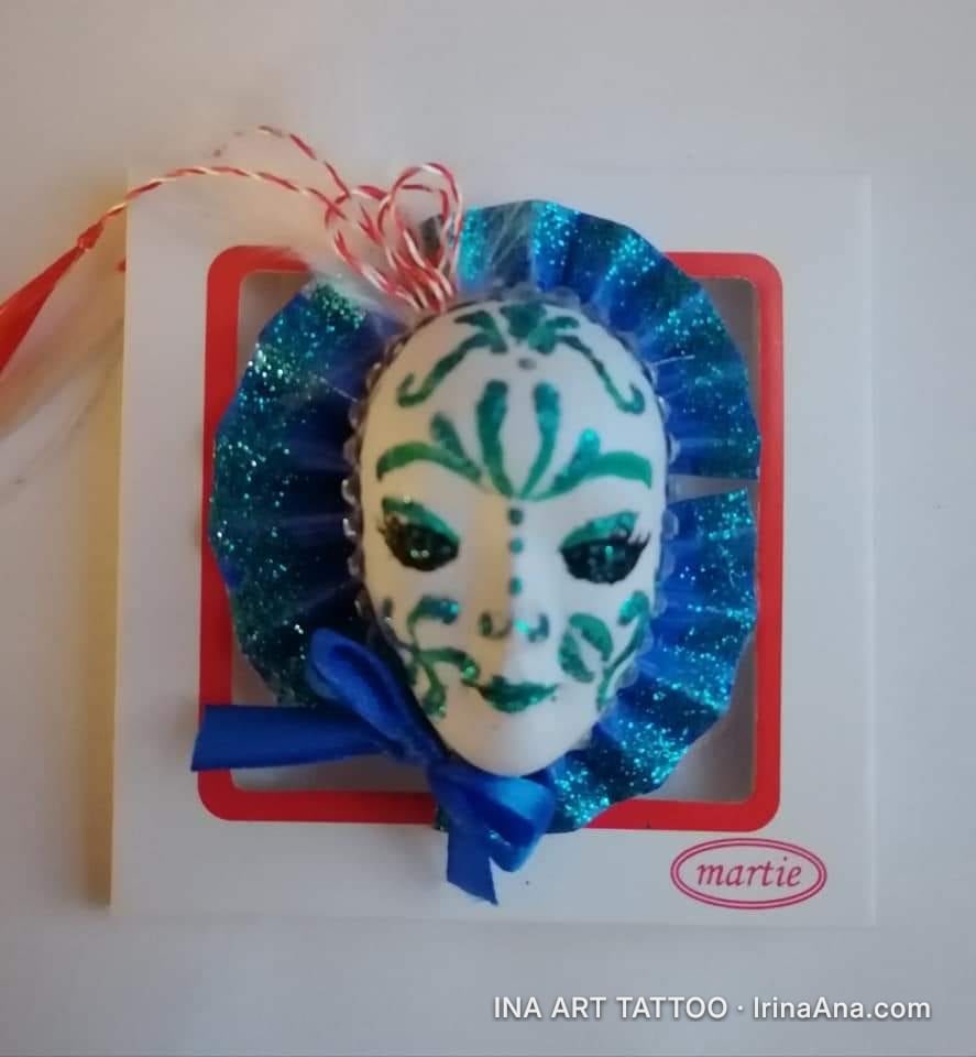 A mărțișor featuring a white mask decorated with green patterns, surrounded by a glittery blue ruffle and a blue bow.