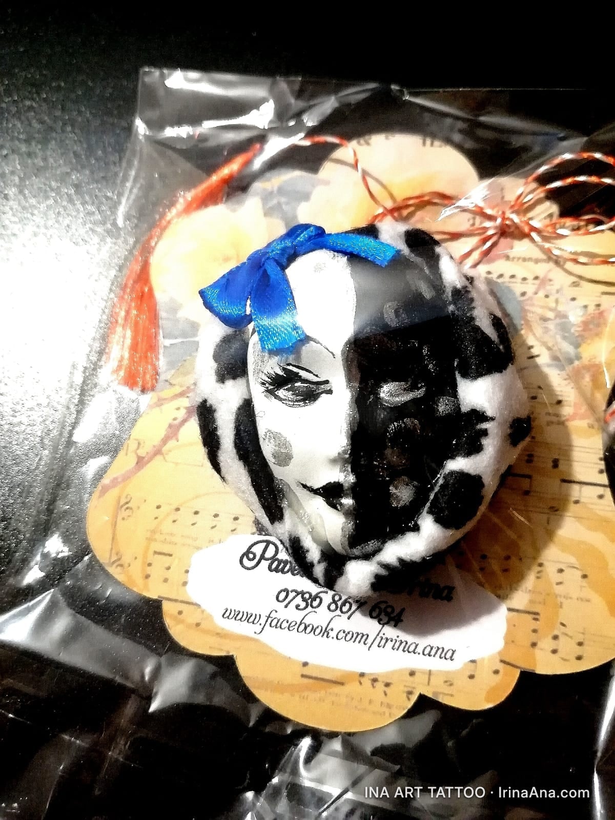 A black and white mask mărțișor with a blue bow, packaged in a transparent bag, on a decorative backing.