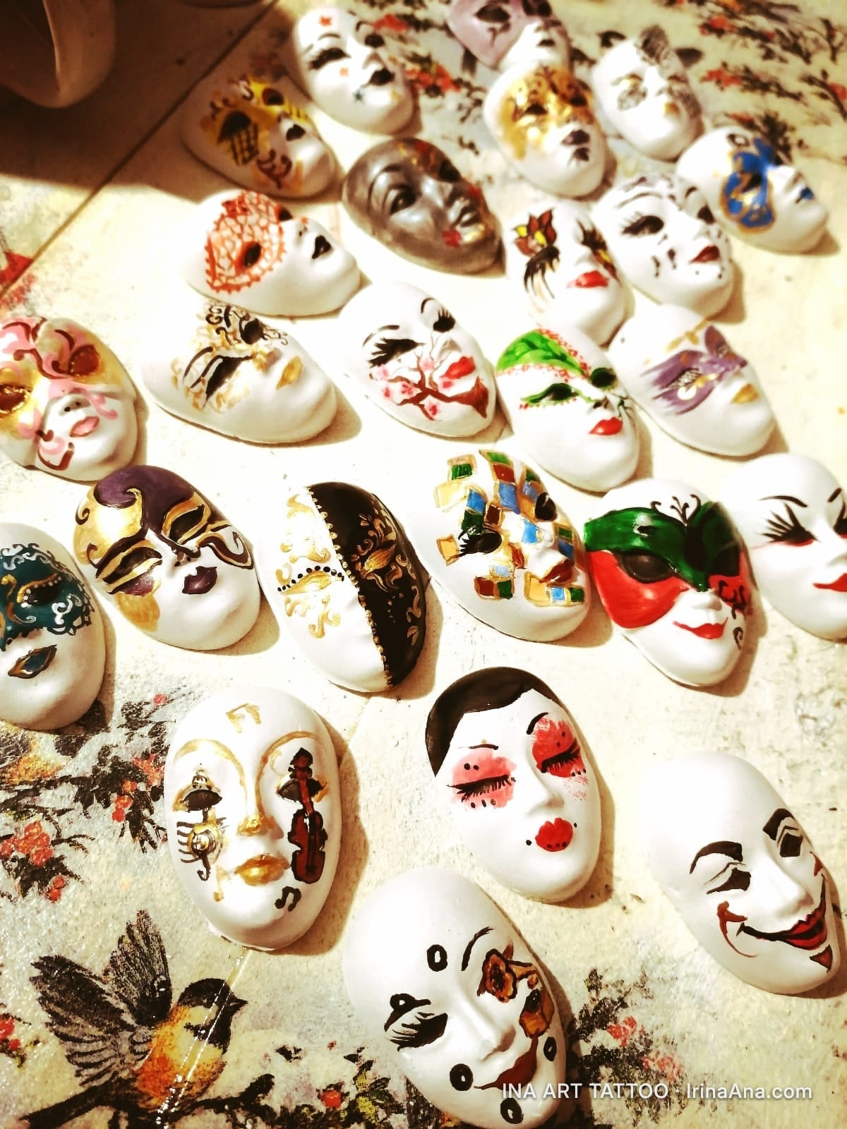 Many small decorative white masks with diverse paintings, arranged on a light-colored surface.