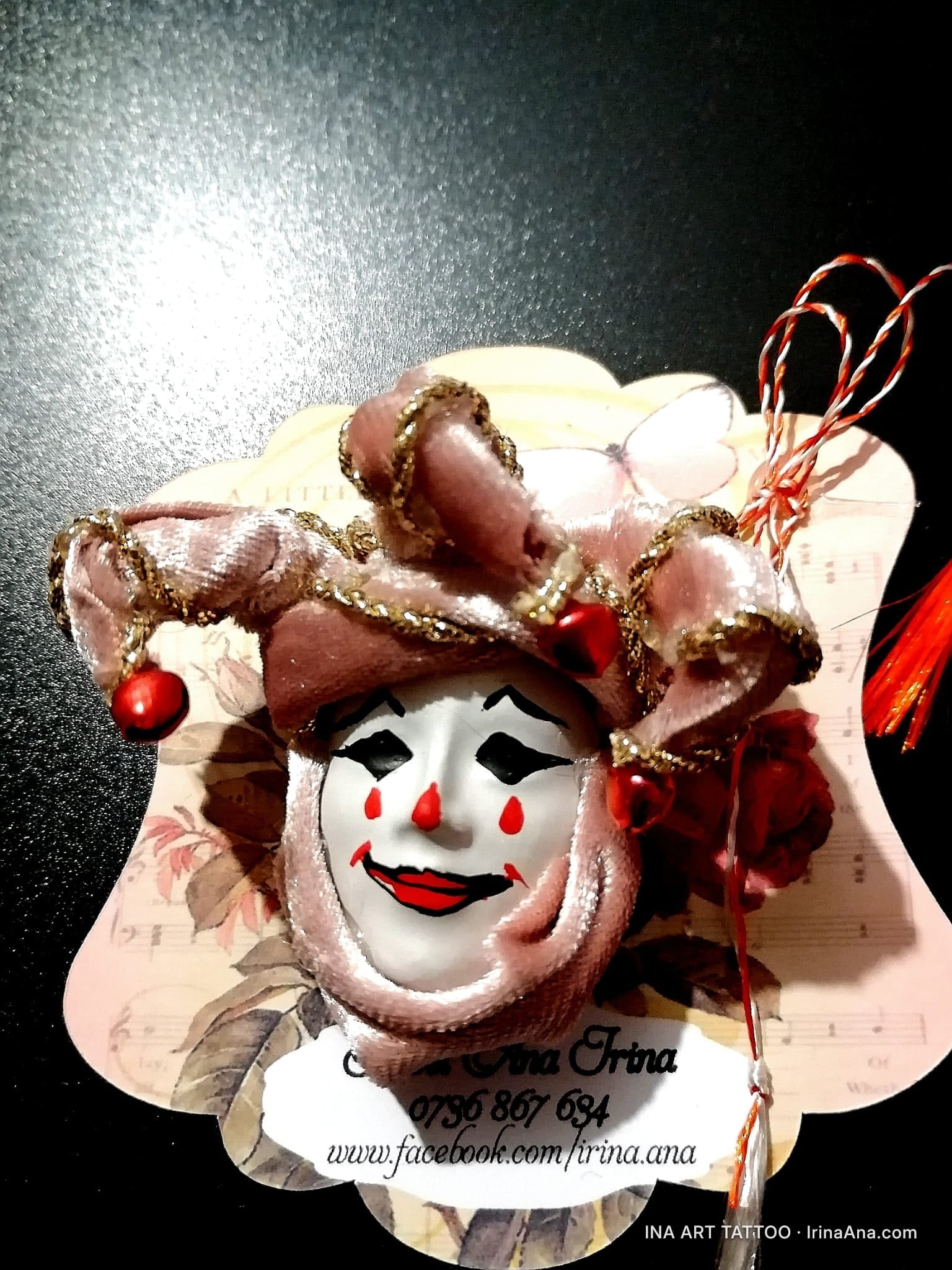 A mărțișor in the shape of a sad clown mask, with red tears, a velvet pink hat, and red tassels.