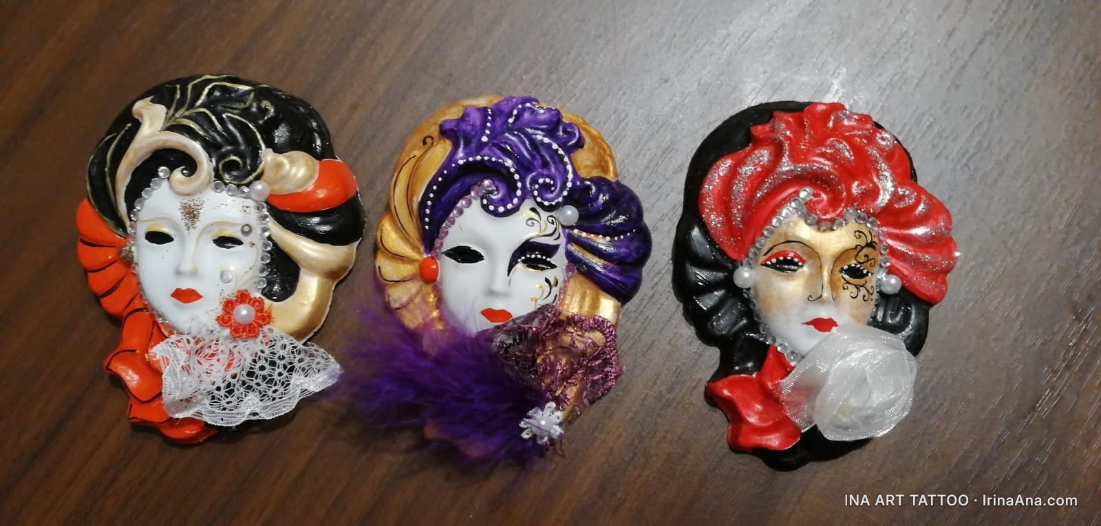 Three Venetian carnival masks, decorated with feathers, lace, and jewels, placed on a wooden surface.
