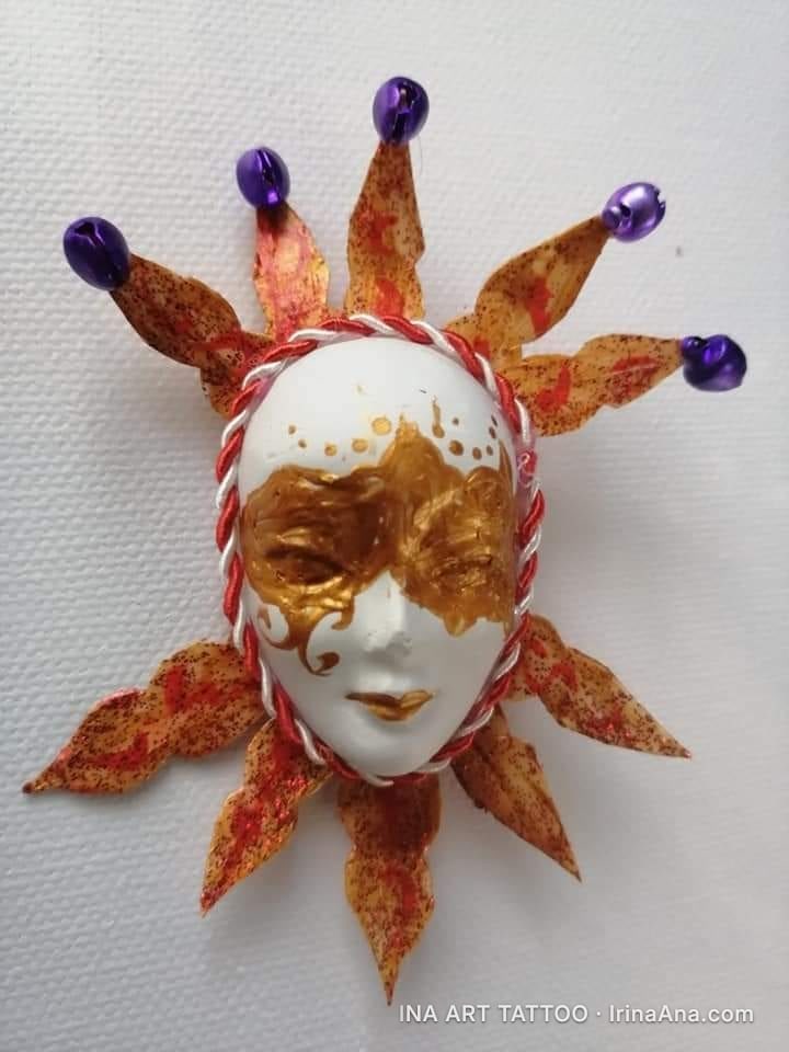 A mărțișor: Venetian jester mask, white and gold, surrounded by orange rays with purple bells.