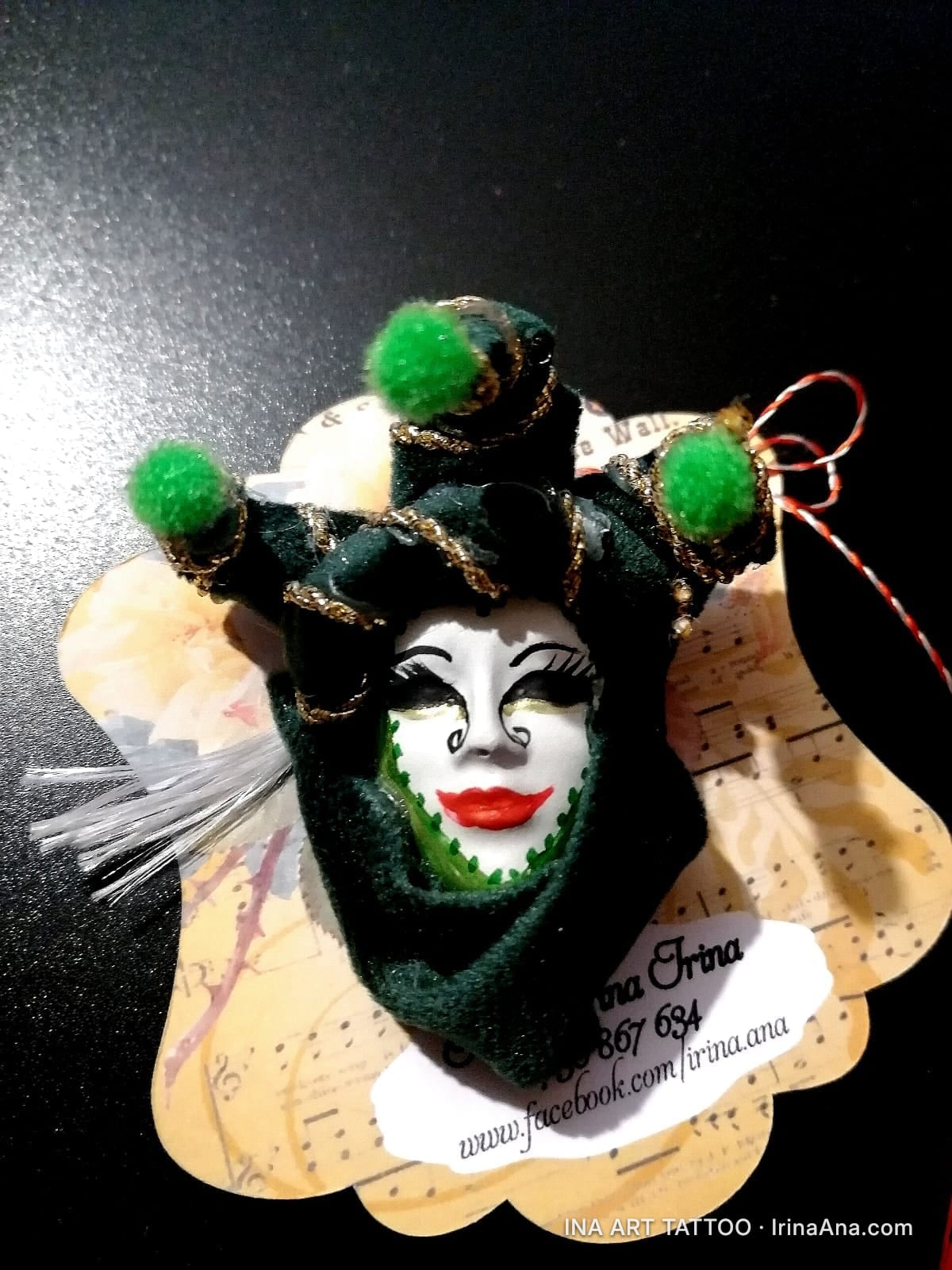 Martisor in the form of a Venetian jester mask, with a white face, red lips, and a green hood with pom-poms.