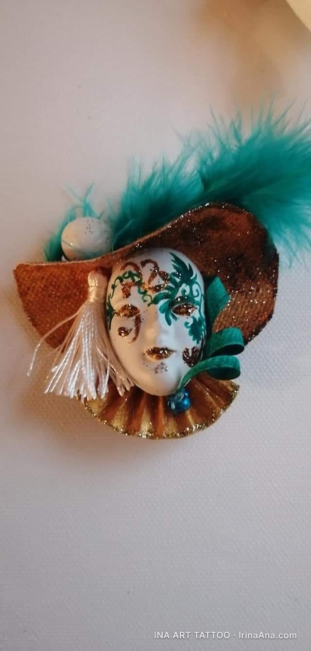 Hand-made craft object: a white Venetian mask with green and gold decorations, wearing a brown hat with turquoise feathers.
