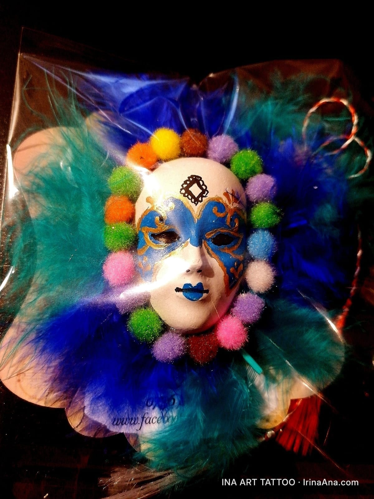 A white Venetian mask with blue and gold details, surrounded by blue and turquoise feathers and colorful pompoms.