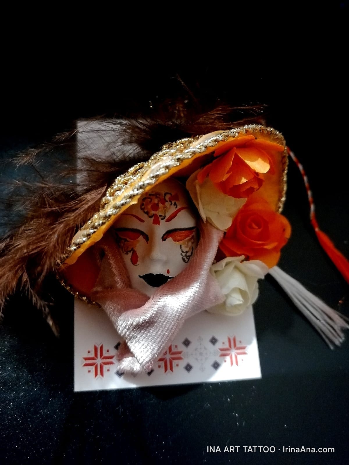 Mărțișor: white Venetian mask with red and black makeup, golden hat with orange flowers and brown feathers, on black backgro…
