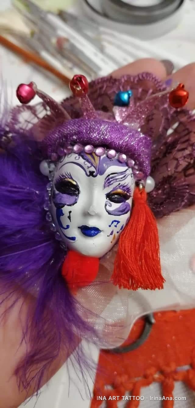 Martisor brooch shaped like a white Venetian mask, with purple decorations, feathers, and red tassels.