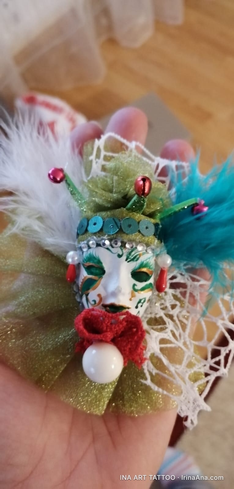 A Venetian mask brooch martisor, decorated with white and turquoise feathers, sequins, and a red bow.